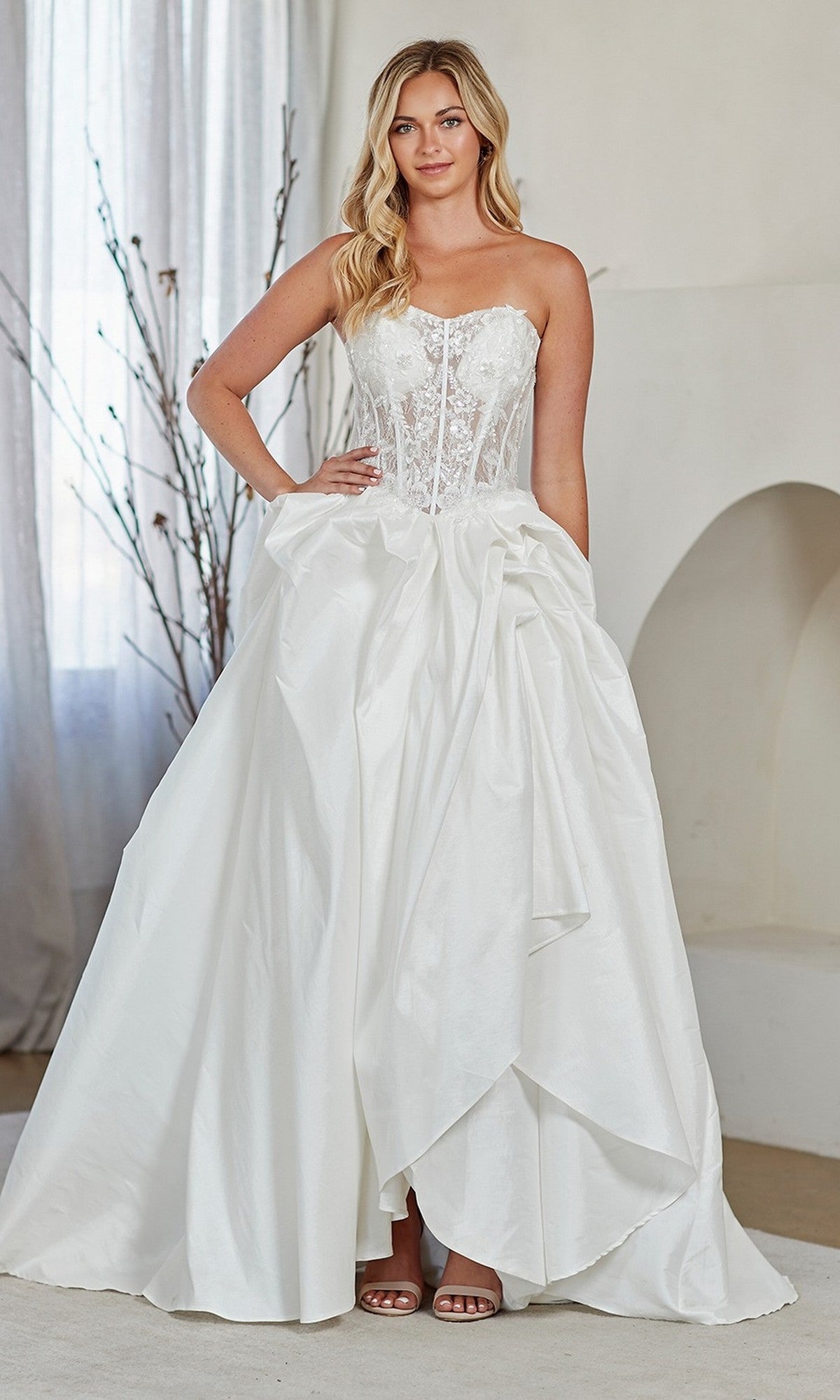 A woman stands indoors on a light floor wearing the Amelia Couture 2085 Long Prom Dress, featuring a strapless white ball gown style with a fitted lace corset bodice and full draped skirt.